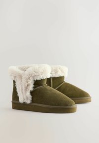 Khaki semišové kotníkové boty s umělou kožešinou, zaoblená špička, plochá podrážka a křížové prošívání. Měkká textura a útulný vzhled.