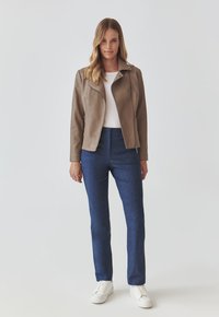 Tan faux leather jacket with a structured collar, paired with dark blue denim pants and white sneakers, featuring a front zip and two side pockets.