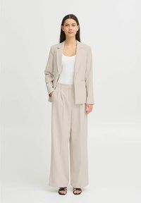 Beige op maat gemaakt pak met een enkelvoudige blazer en wijde broekspijpen. De stof is glad met een lichte glans; heeft een zak en een minimalistisch ontwerp.