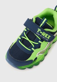 Plava i zelena dječja tenisica s mrežastim materijalom, Velcro trakom i teksturiranim đonom. Sadrži grafiku T-Rexa i žive naglaske.