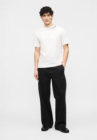 Νεαρός άνδρας με κατσαρά μαλλιά που φοράει λευκό T-shirt με το λογότυπο EA7, μαύρο παντελόνι με φαρδιά μπατζάκια και μαύρα αθλητικά παπούτσια, στέκεται με το ένα χέρι στην τσέπη.