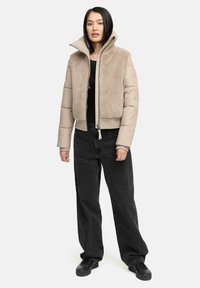 Beige pufferjack met een getextureerde, zachte voorkant, hoge kraag en geribde manchetten. Draag het over een zwarte top in combinatie met zwarte wijde broek en zwarte schoenen.