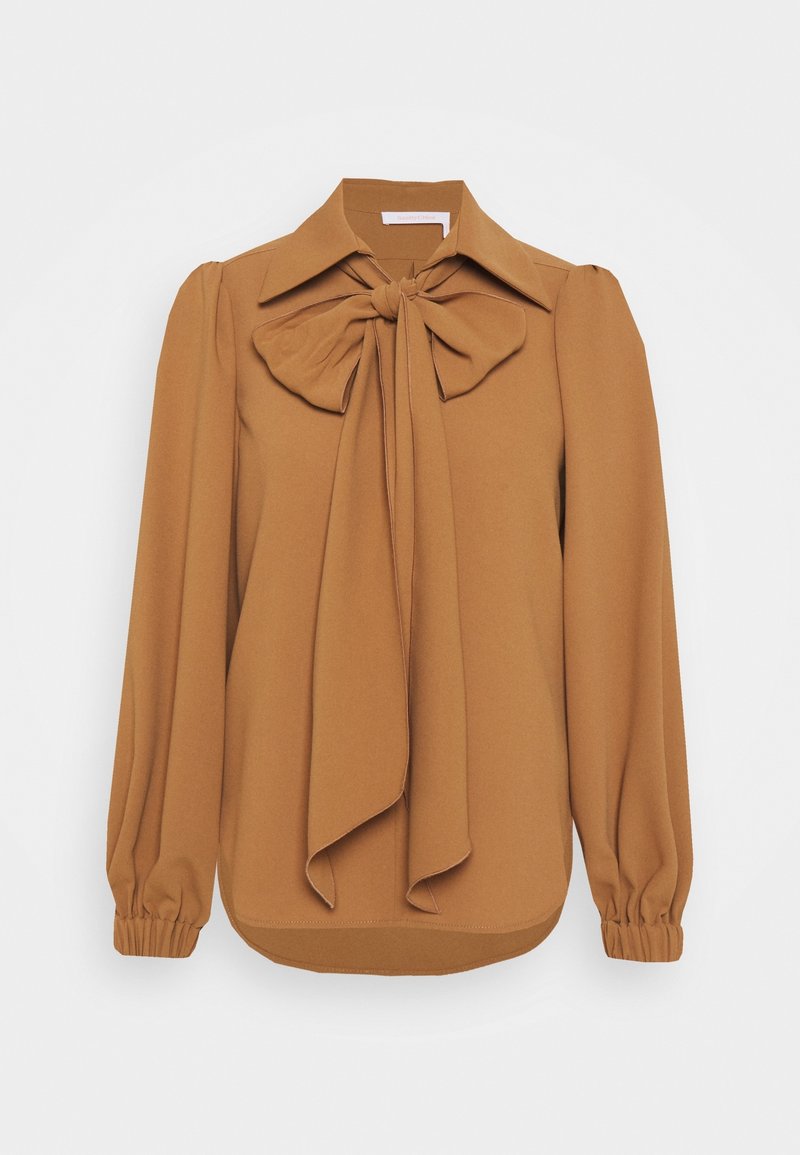 Blouse camel à manches longues avec un grand nœud papillon à l’avant, poignets froncés et col pointu sur fond blanc.
