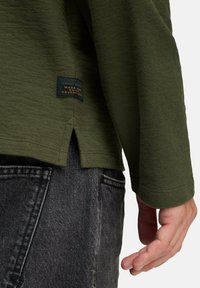 Close-up van een persoon die een olijfgroene lange mouwen shirt draagt met een zij-split en een klein zwart label nabij de zoom, gecombineerd met donkergrijze jeans.