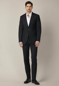 Czarny garnitur o slim fit, z wyłogowanym kołnierzykiem, jednorzędowym kroju i dwoma przednimi kieszeniami. Biała koszula pod spodem, w zestawie z czarnymi butami.