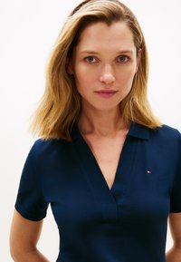 Tengerészkék galléros póló V-nyakú kialakítással, rövid ujjakkal, és egy kis piros-fehér logóval a bal oldalon.