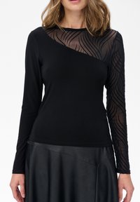 Schwarzes Langarmshirt mit einem transparenten Mesh-Einsatz mit gewellten Mustern auf einer Seite. Der glatte Stoff und das eng anliegende Design ergänzen einen schwarzen Rock.