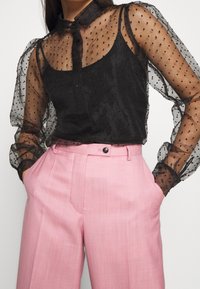 Blouse noire avec manches transparentes à pois, boutonnée sur le devant, associée à un pantalon taille haute rose clair avec une texture lisse et un bouton unique.