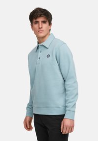 Ljusblå sweatshirt med klassisk krage, knappslå och en liten mörk logotyp på bröstet. Har ribbade muddar och nederkant.