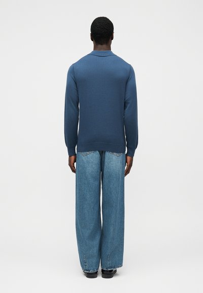 Άνδρας στέκεται με την πλάτη προς το μέρος μας, φορώντας μπλε πουκάμισο μακρυμάνικο, ανοιχτόχρωμα τζιν με φαρδιά πόδια και μαύρα παπούτσια, μπροστά σε απλό γκρι φόντο.