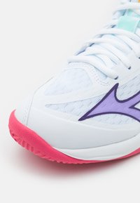 Bílé sportovní boty s texturovaným svrškem, detail v podobě fialového loga, růžovou podrážkou a bílými tkaničkami. Detailní záběr zachycuje tvar a design.