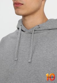 šedá mikina s kapucí a stahovací šňůrkou, s jemnou texturou. Detailní pohled ukazuje výstřih a švy.