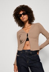 Light brown ribbed cardigan with a v-neck and front clasp, worn over a black top, paired with dark blue jeans. Model has curly hair and sunglasses.