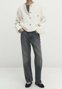 Kremowa kurtka typu button-up o teksturze, z dwiema przednimi kieszeniami, zestawiona z szarymi dżinsami prostymi i czarnymi butami. Minimalistyczny design.