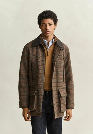 Jonge man die een bruine geruite jas draagt over een beige trui met rits en een lichtblauw overhemd met kraag, tegen een effen beige achtergrond.