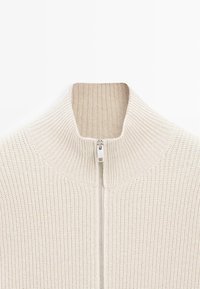 Maglione lavorato a maglia di colore crema con colletto alto e zip a lunghezza completa. Presenta una texture a coste e un design aderente, offrendo un aspetto accogliente.