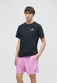 Νεαρός άνδρας που φοράει μαύρο μπλουζάκι Nike και ροζ σορτς Nike, στέκεται με το ένα χέρι στην τσέπη μπροστά σε απλό ανοιχτό γκρι φόντο.