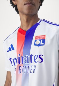 Bílý fotbalový dres s červenými a modrými diagonálními pruhy, obsahující logo "Emirates", znak "Olympique Lyonnais" a texturovanou tkaninu.
