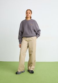 Huppari harmaana, ylipitkä ja brodeeratulla logolla, beige suorat housut ja mustat harmailla yksityiskohdilla varustetut lenkkarit, vaaleaa taustaa vasten.