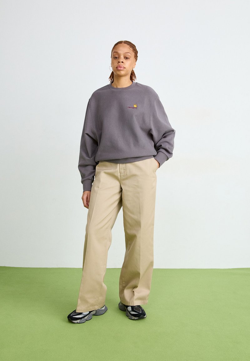 Grå oversized sweatshirt med broderad logotyp, beige raka byxor och svarta sneakers med grå accenter, mot en ljus bakgrund.