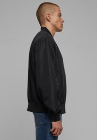 Veste bombardier noire avec une texture lisse, col et cuffs côtelés, et poches latérales, présentée sur une personne de profil.