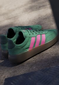 Baskets vertes Adidas avec des bandes roses et des semelles en gomme placées sur une surface en béton ensoleillée.