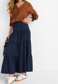 Cardigan marrone con scollo a V, abbinato a una gonna maxi blu navy a più strati. Sneakers bianche con accenti bordeaux e design a stelle dorate.