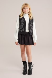 Črn vezeni brezrokavnik preko bele turtleneck majice, v kombinaciji s črnimi kratkimi hlačami in črnimi škornji do kolen z okrasnimi zaponkami.