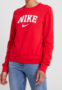 Röd sweatshirt i bomullsblandning med en vit "NIKE"-logga och swoosh. Ribbstickade muddar och fåll ger textur och struktur.