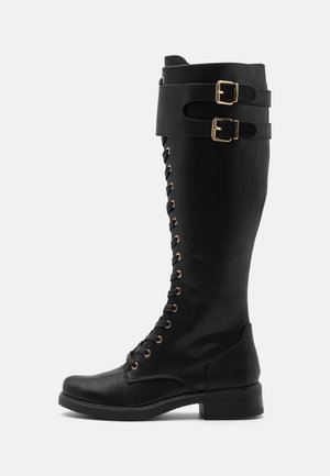 Botte noire en cuir montant jusqu'au genou avec lacets sur le devant et deux brides à boucle dorée près du haut, semelle plate et petit talon.