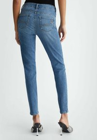 Jeans de mezclilla azul de corte ajustado y altura media con bolsillos traseros que presentan un bordado sutil; llevados con tacones destalonados negros sobre un fondo liso.