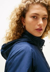 Granatowa kurtka z kapturem, wykonana z teksturowanej, lekkiej tkaniny. Posiada sznurek przy dekolcie oraz widoczne detale zamka.