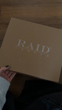 Brun pappeske med hvittrykt logo "RAID LONDON." Eskens hjørne er litt åpent, og avslører en strukturert overflate.