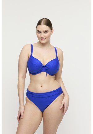 Vrouw met een lichte huid en haar in een knot, gekleed in een blauw, getextureerd bikini, staand tegen een effen lichte achtergrond.