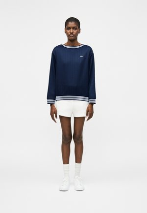 Femme debout portant un haut à manches longues bleu marine avec des poignets et un col blancs rayés, un short blanc, des chaussettes blanches et des baskets blanches.