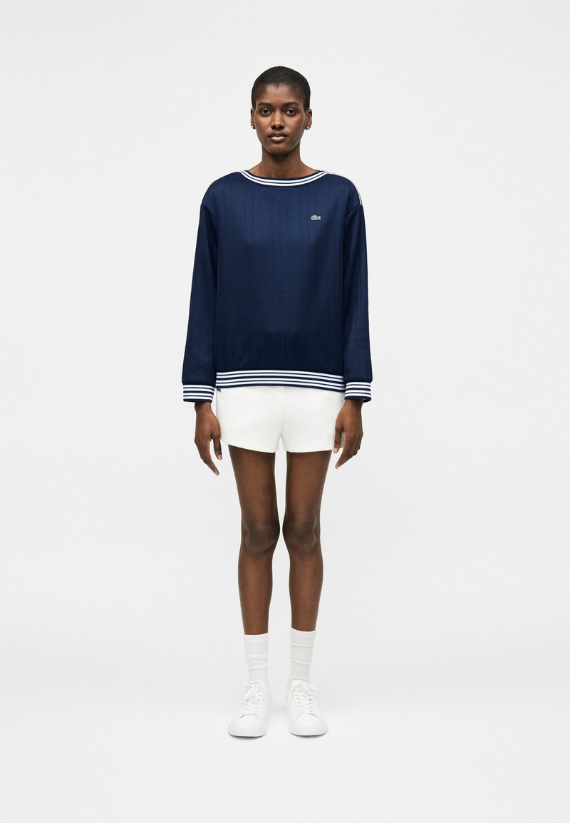 Femme debout portant un haut à manches longues bleu marine avec des poignets et un col blancs rayés, un short blanc, des chaussettes blanches et des baskets blanches.
