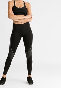 Žena stojící s překříženýma rukama, oblečená v černém sportovním topu, černých legínách se šedými panely a černých sportovních botách.