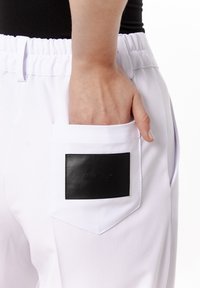 Białe spodnie z elastycznym pasem, z tylną kieszenią, w której znajduje się czarna prostokątna naszywka z napisem logo. Gładka faktura materiału.