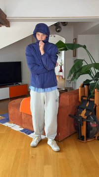 Personne portant une veste à capuche bleue, un pantalon de survêtement gris clair et des baskets blanches, debout à l'intérieur près d'un canapé orange et d'une grande plante verte.