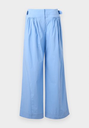 Kvinde står med hænderne i lommerne, iført en hvid skjorte med knapper, beige top nedenunder, brede lyseblå bukser og hvide spidse sko.