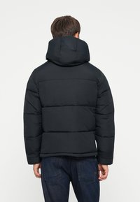 Sort puffjakke med hætte, der har horisontale syninger og en afslappet pasform. Båret over mørk denim, vist fra ryggen.