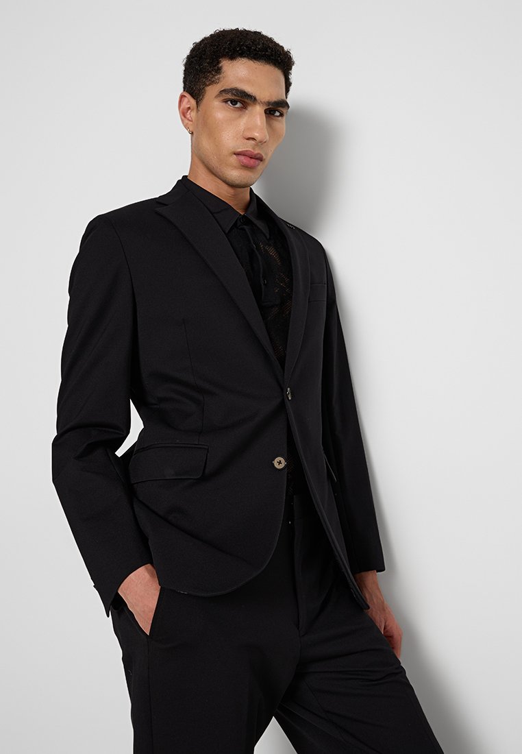 Un modello posa su uno sfondo bianco, indossando una giacca di abito nera su una camicia nera, mostrando un'espressione sicura di sé.