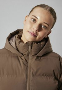 Chaqueta acolchada con capucha de color marrón, con cierre de cremallera y cuello de canalé. La tela parece suave y ligeramente brillante, con una textura suave.