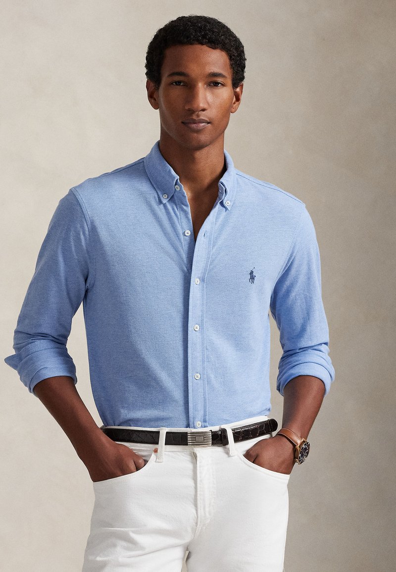 Ανοιχτό μπλε πουκάμισο με κουμπιά από μαλακό ύφα�σμα, μακριά μανίκια διπλωμένα στους καρπούς, με μικρό κεντημένο λογότυπο στο στήθος. Φοριέται με λευκά παντελόνια.