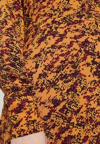 Maglietta a manica lunga in tessuto arancione con un motivo testurizzato di macchie nere e bordeaux. Materiale morbido con un design dal fit rilassato.