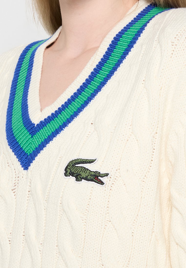 Maglione a trecce crema con scollo a V, dettagli in costina blu e verde e logo di coccodrillo verde sul lato sinistro del petto.
