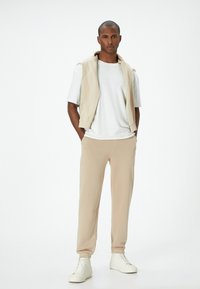 Felpa senza maniche beige sopra una maglietta bianca, abbinata a pantaloni jogger beige e sneakers bianche. Design semplice, texture morbida, vestibilità rilassata.