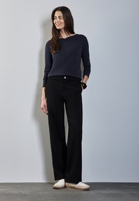 Musta leveälahkeinen housu, jossa on sileä rakenne ja nappiyksityiskohta vyötäröllä, yhdistettynä tummansiniseen pitkähihaiseen paitaan ja vaaleanvärisiin kenkiin.