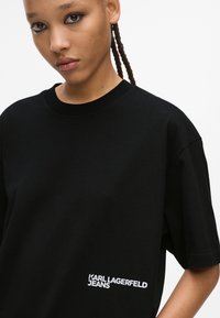Sort bomulds t-shirt med rund halsudskæring og korte ærmer. Har en hvid logo tekst "KARL LAGERFELD JEANS" nederst til venstre.