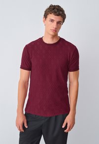 Burgundy short-sleeve t-shirt featuring textured diamond pattern, crew neck, and contrasting trim on sleeves. Worn with dark pants.